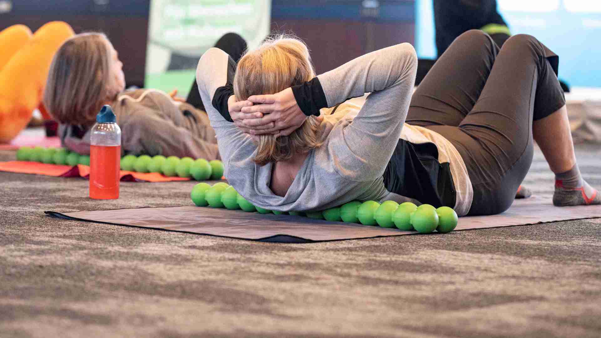 gesund_leben_messe_Fitnesskurs Zwei Besucherinnen liegen auf dem Rücken und nehmen aktiv an einem Fitnessprogramm teil, während sie gemeinsam eine Übung nachmachen.